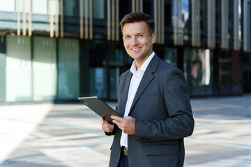 A confident businessman smiling while holding a tablet in front of a modern office building.