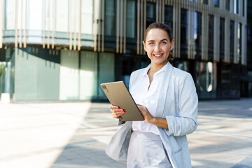 A confident businesswoman holding a tablet, smiling brightly in front of a modern glass office building.