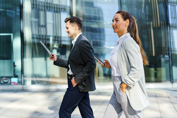 Two business professionals walking side by side outside a modern office building, engaged with a tablet and smartphone.