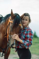 Happy blonde with horse in forest. Woman and a horse walking through the field during the day. Dressed in a plaid shirt and black leggings.