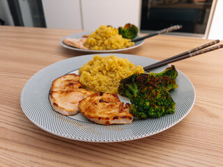 Two plates with grilled chicken, quinoa, and broccoli, one in focus and the other in the background, served with chopsticks on a wooden table.