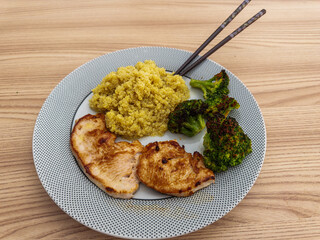 A healthy meal of grilled chicken breast, quinoa, and roasted broccoli served with chopsticks on a dotted plate, set on a wooden table.