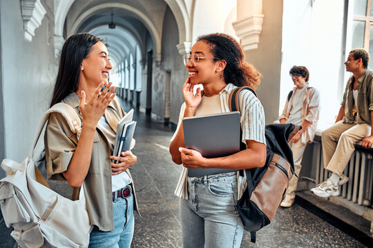 Two sincere college students are chatting while standing in the hallway. Cheerful female student friends are talking during a break.