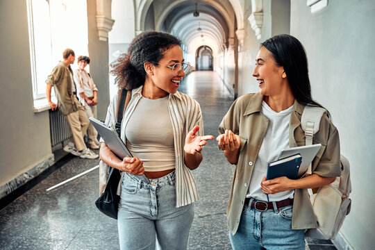  Modern stylish beautiful sincere female students holding books and study gadgets in their hands and walking down the corridor of the university. Concept of education and student life. - Powered by Adobe