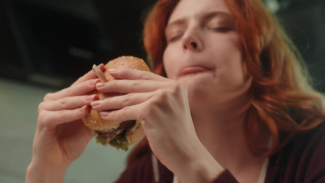 Happy hungry Caucasian woman female girl client customer eat chew tasty burger eating delicious juicy hamburger enjoy chewing cheeseburger indoors kitchen room. Fast food delivery unhealthy nutrition