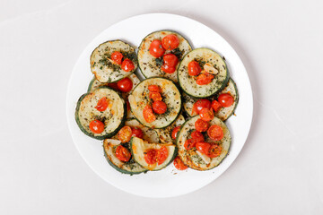 Fried zucchini with garlic and cherry tomatoes on white plate and grey marble table.