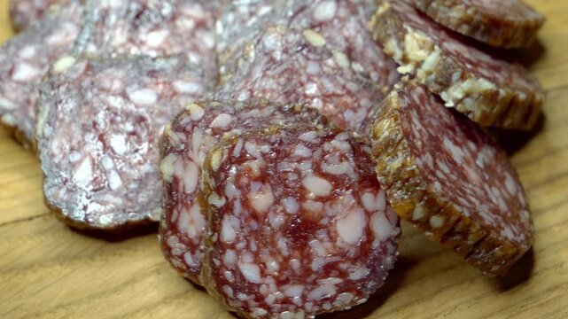 Several slices of salami are arranged on a wooden surface. The salami is dark red with white specks, and has a rough texture. A close-up view of salami slices on a wooden cutting board.