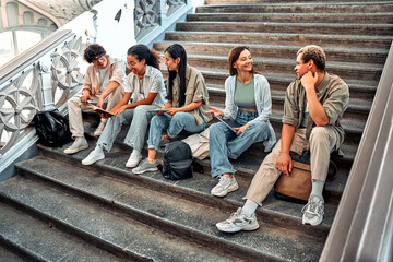 Fototapeta premium A group of student friends are sitting on the steps of the university inside and talking, getting ready for classes. Modern students.
