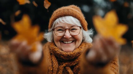 A joyful elderly woman in warm clothing, smiling and holding autumn leaves, conveys a cheerful and happy autumn feeling, with a background of falling leaves and nature.
