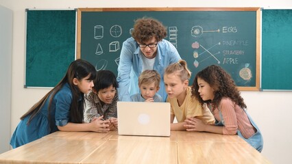 Caucasian teacher and diverse student looking at laptop at classroom. Cute multicultural children learning about coding program or generated program by using software while teacher teaching. Pedagogy.