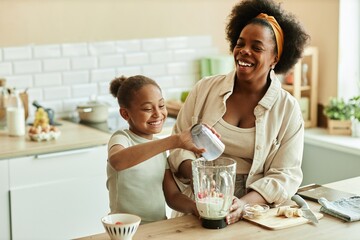 Laughing African American young mother and little daughter enjoying quality time while cooking together making fruit smoothie in blender at beige kitchen, copy space
