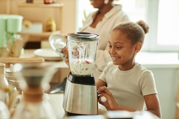 Smiling African American little girl with ponytail hair cheerfully looking at blender mixing ingredients for fruit smoothie while making healthy dessert at sunlit kitchen, copy space