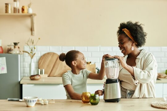 Side view of smiling African American mom and little daughter having fun while cooking breakfast together making smoothie in blender on large wooden counter at cozy beige kitchen, copy space