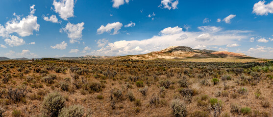 Fossil Butte National Monument in Wyoming, the vibrant blue sky and white clouds contrast with the orange hills and green grass, while rugged rock formations and sparse shrubs across the horizon.