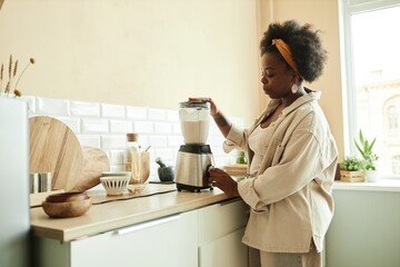 Side view of curly Black woman using food blender mixing ingredients for milk smoothie while cooking healthy breakfast at beige sunlit kitchen, copy space