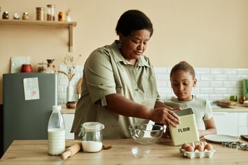 Medium shot of caring African American grandmother taking metal flour bin teaching curious little grandchild how to cook pancakes at cozy beige kitchen with large wooden counter, copy space