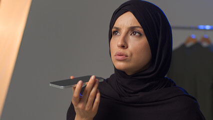 Angry displeased Islamic Muslim Arabian woman model lady speaking talk loudspeaker negotiate mobile phone smartphone call. Annoyed girl in hijab female record audio voice message voicemail in studio