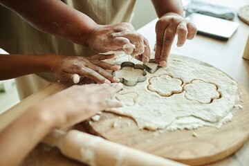 Close up on hands of African American woman and young girl cutting flower shaped cookies from dough with metal cookie cutters while making homemade biscuits at kitchen, copy space