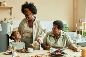 Smiling little girl with loving mother and granny having delicious cake sitting at table while mom...