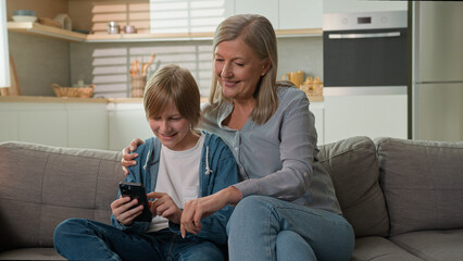 Happy elderly mature Caucasian woman senior female granny mother smile hug grandson kid little boy child typing screen mobile phone play online choosing goods browse Internet together family home sofa