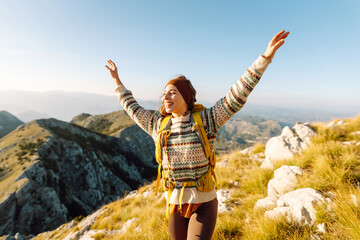 Cheerful young woman sits on a grassy slope, smiling as the sun sets behind the mountains. She has a backpack beside her and is taking a moment to enjoy the beautiful landscape. Lifestyle, tourism.