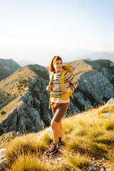 Obraz premium Cheerful young woman sits on a grassy slope, smiling as the sun sets behind the mountains. She has a backpack beside her and is taking a moment to enjoy the beautiful landscape. Lifestyle, tourism.