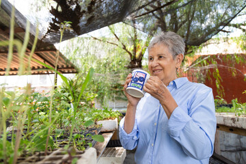 A woman is holding a mug and smiling while looking at plants. The plants are in pots and are growing in a greenhouse