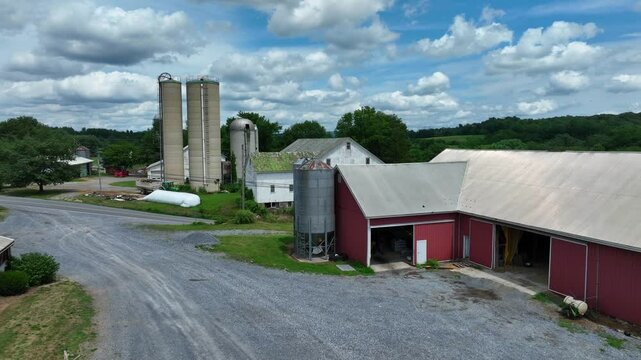 Farmer walking on path of own farmstead with silo storage, barn and stables in USA. Sunny day with clouds in summer. Aerial approaching shot.