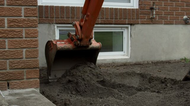 Mini excavator carefully excavating foundation of residential home - close up of bucket in front of basement window