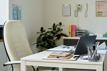 Clean and organized white desk featuring notebooks, pens, coffee cup, and open laptop in modern office environment. Chair and green plant adding touch of nature