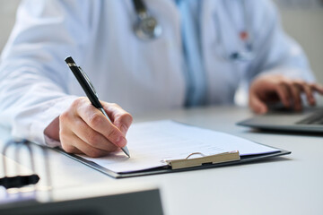 Fototapeta premium Medical professional writing on clipboard with pen while utilizing laptop at desk in bright office, half-closed blinds in background, stethoscope around neck