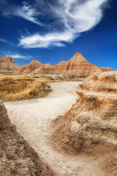 Sandy path leads through the striated peaks to the higher peaks in the background stark against a blue sky.