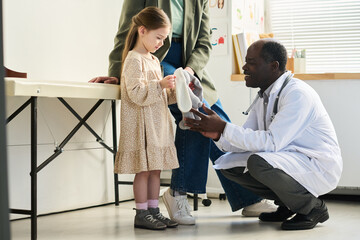 Senior doctor kneeling and assisting young girl holding white stuffed animal in medical office environment with diverse people present and medical equipment surrounding