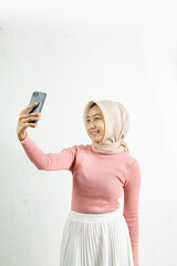 Naklejka premium Advertisement The Muslim teenager took a selfie with a cellphone with a white background. The woman was wearing a hijab and a pink woven dress and white skirt.