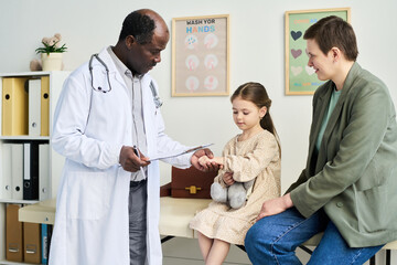Fototapeta premium Pediatrician consulting with child and mother during medical exam. Doctor wearing white coat and checking medical notes while sitting on examination table