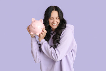 Pretty young woman with piggy bank on lilac background