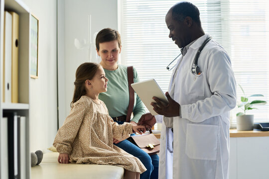 Engaging with child patient and parent while holding tablet in office. Medical professional smiling, creating comforting environment for young patient, promoting positive interaction