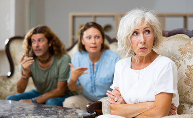 Dissatisfied mother-in-law siting on a chair against of a confused husband and wife on the sofa in...