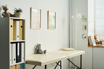 Pediatric examination room with minimalist decor, featuring soft toys on shelves, framed pictures, an examination table, and medical equipment against white walls creating an inviting atmosphere