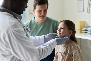 Doctor in white coat carefully examining young girl throat, while mother watches attentively seated beside her in a well-lit clinics