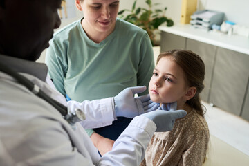 Obraz premium Doctor checking young girl's throat in clinic with mother sitting beside Medical professional wearing gloves and stethoscope