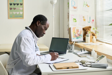 Fototapeta premium African American doctor writing notes at desk in office with medical equipment and laptop visible. Office is decorated with educational posters and children's drawings adding warmth
