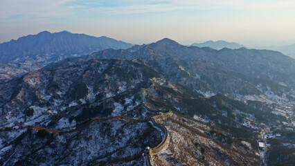 The Great Wall of China running through snow-covered, tree-lined mountains under a partly cloudy sky.
