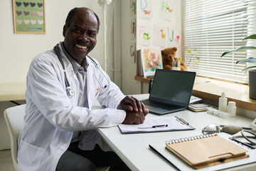 Smiling African American doctor sitting in office surrounded by medical equipment, holding clipboard and reviewing documents while looking content and approachable