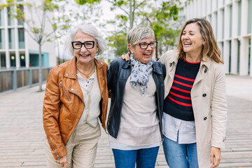 Three mature retired women laughing while walking together outside. Small group of 60s females having fun enjoying a conversation bonding at city street. Elderly friendship concept.