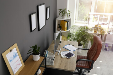 Interior of office with workplace, armchair, modern computer, houseplant and shelving unit near black wall