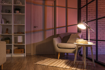 Comfortable armchair, table and glowing lamp in interior of living room