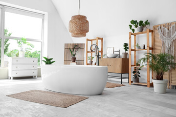 Bathtub, wooden shelving unit, chest of drawers and houseplants in interior of light bathroom