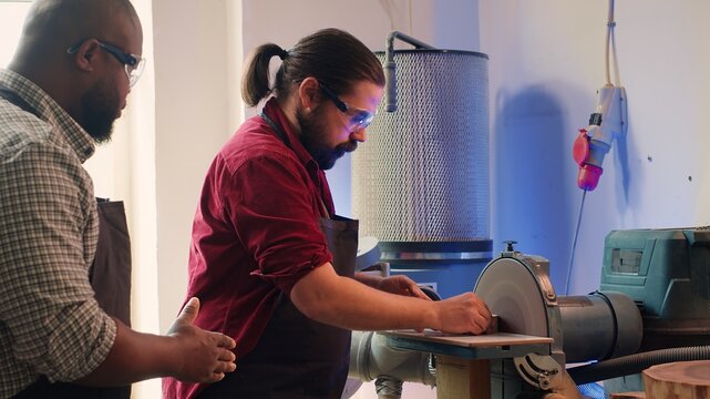 Man teaching african american trainee how to shape wooden bowl on disc sander in assembling shop. Worker learning from woodworking professional how to use grinding machine rotating disc, camera A