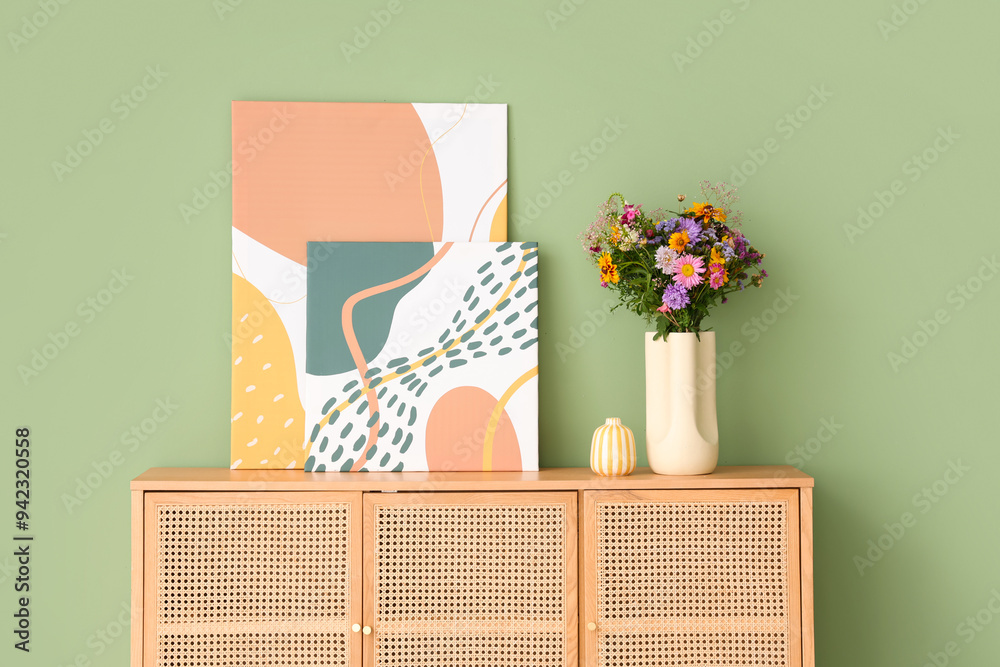 Wall mural pictures and vase with wild flowers on chest of drawers near green wall in room